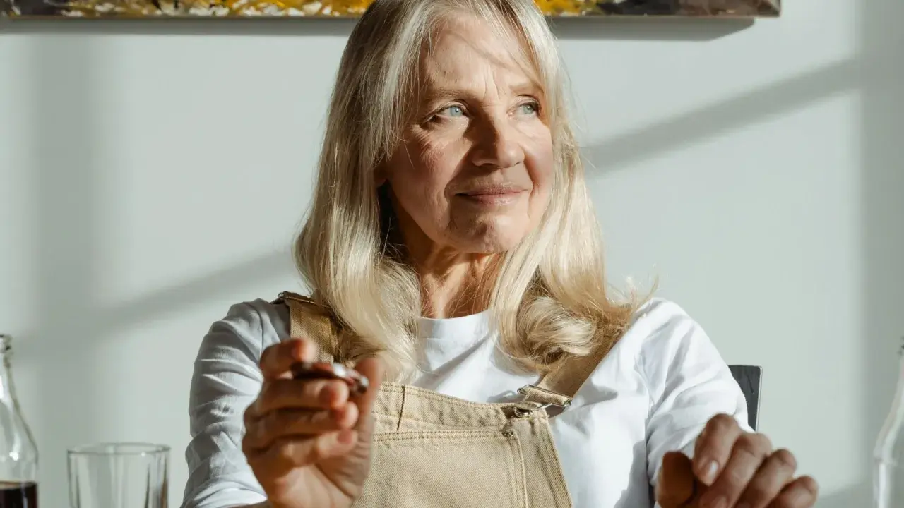 Donna anziana con capelli biondi e occhi azzurri, le rughe sul viso raccontano storie di vita. Tiene in mano un oggetto.
