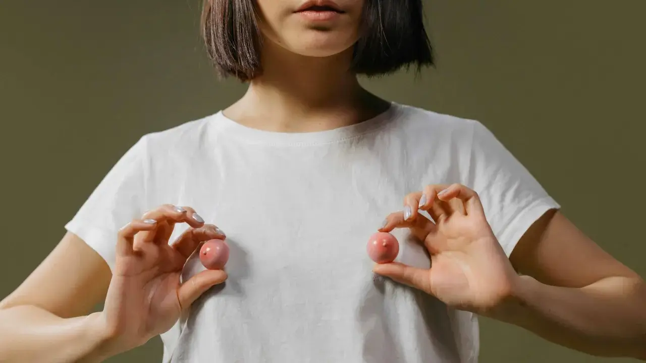 Mani che tengono due sfere rosa, una che ricorda un capezzolo e areola, su una maglietta bianca.