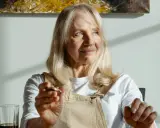 Donna anziana con capelli biondi e occhi azzurri, le rughe sul viso raccontano storie di vita. Tiene in mano un oggetto.