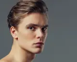 Primo piano di un giovane uomo con capelli biondi pettinati all'indietro e occhi azzurri penetranti. Il suo viso, con un leggero tocco di filler uomo viso, appare definito e curato.