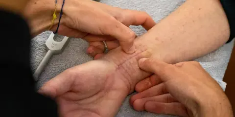 Mani esperte che mostrano come massaggiare una cicatrice sul polso, con un diapason in primo piano.