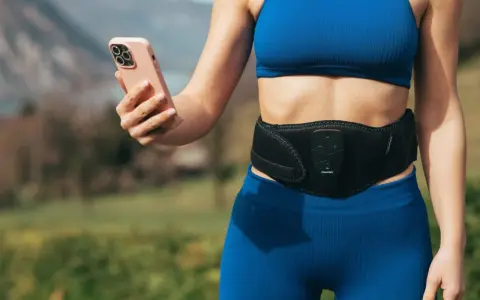 Donna con fascia addominale per tonificare la pancia molle dopo parto, mentre controlla l'app sul suo smartphone.