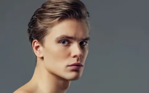 Primo piano di un giovane uomo con capelli biondi pettinati all'indietro e occhi azzurri penetranti. Il suo viso, con un leggero tocco di filler uomo viso, appare definito e curato.