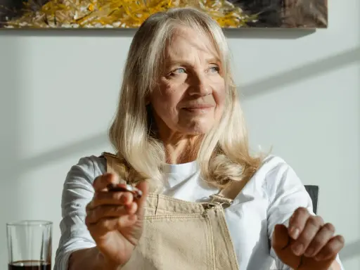 Donna anziana con capelli biondi e occhi azzurri, le rughe sul viso raccontano storie di vita. Tiene in mano un oggetto.
